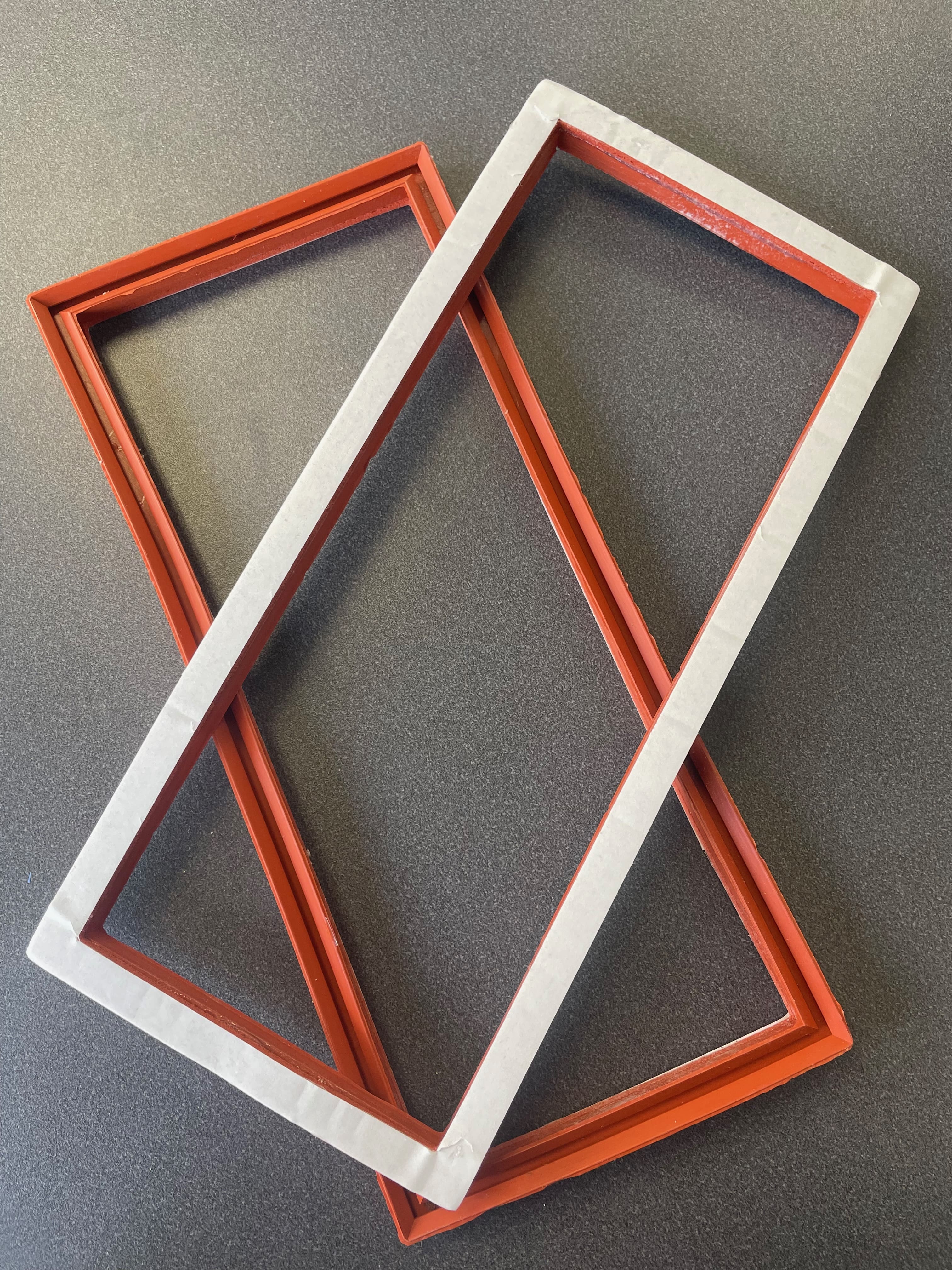 Two rectangular red molded rubber parts with adhesive strips on the top. One is sitting on top of the other. The bottom part is upside down.